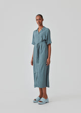Relaxed shirt dress in blue made from EcoVero viscose. DafneMD long dress has a resort collar, button closure in front, tie belt at the waist, and short sleeves. The model is 177 cm and wears a size S/36.