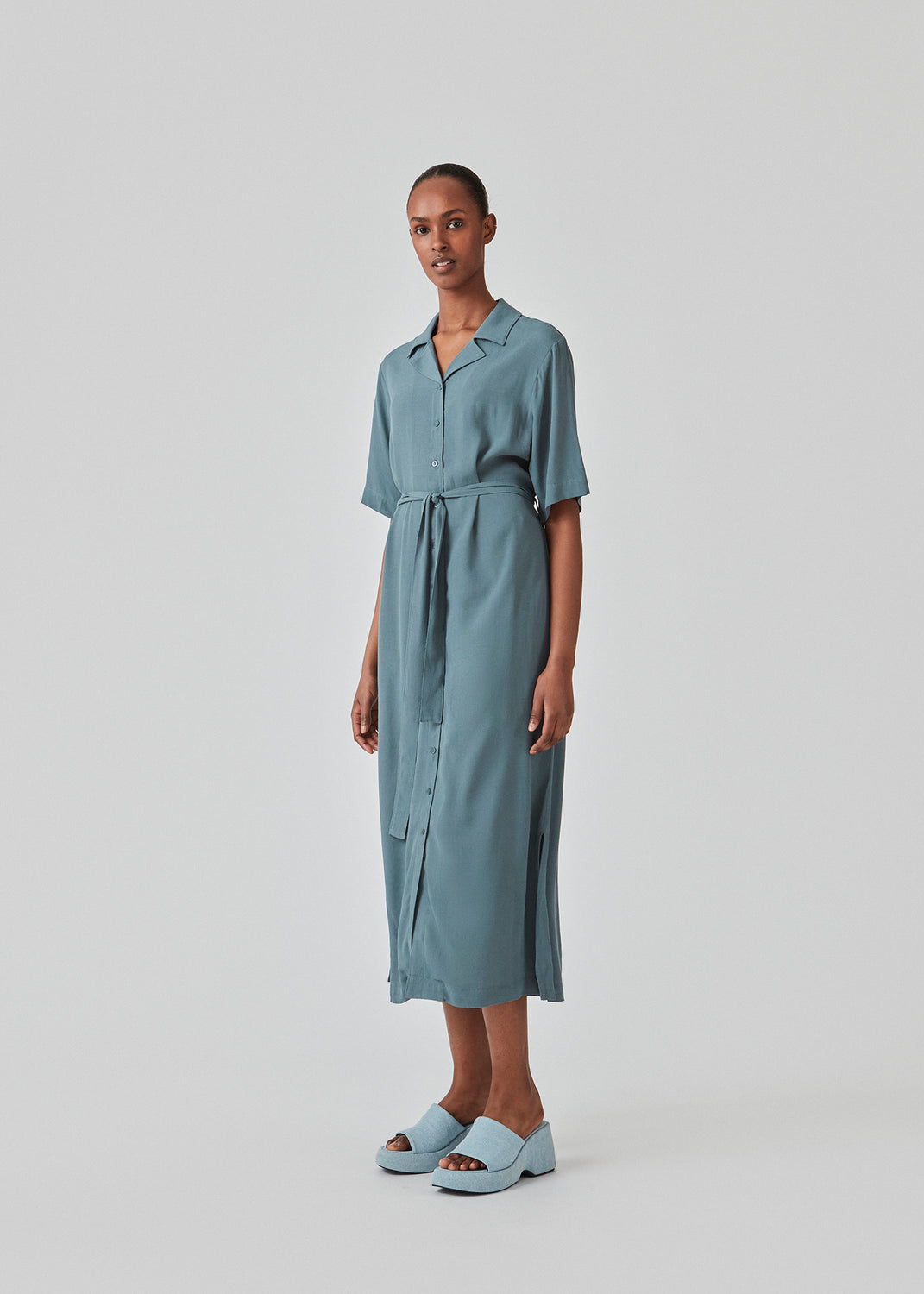 Relaxed shirt dress in blue made from EcoVero viscose. DafneMD long dress has a resort collar, button closure in front, tie belt at the waist, and short sleeves. The model is 177 cm and wears a size S/36.