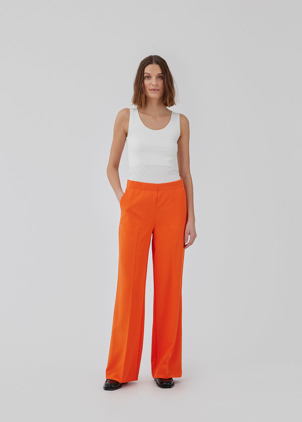 Classic pants in brigt red with pressfolds and straight legs. Nelli pants are closes by an hidden zipper at the side with an elastic waistband for a more comfortable fit. The model is 173 cm and wears a size S/36