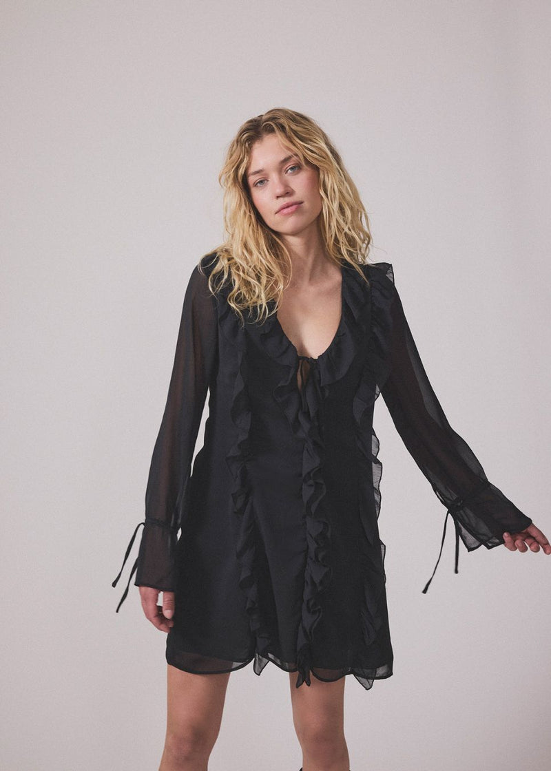 Short black dress with frill details and a loose fit. NorrisMD dress has a v-neck and long, wide sleeves with tiebands.