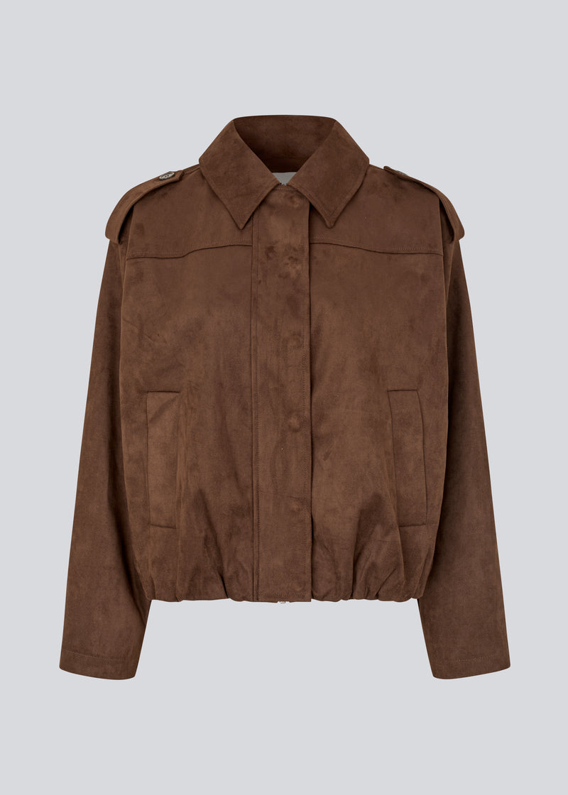 Short brown jacket made from a soft, suede-like material. Features a classic shirt collar, hidden zip and button closure at the front, and practical front pockets. The structured shoulders with strap details and the slightly elasticated hem add a modern, tailored touch. A versatile and stylish piece, perfect for transitional weather and everyday wear.