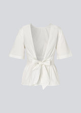 Feminine top with an open back and tie detail. The AikaMD Top features short sleeves, a round neckline, an A-line hem with pleating, and an attached band that cinches at the waist and ties at the back. The top is made from 100% organic cotton.