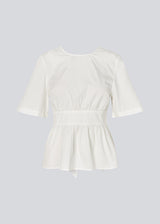 Feminine top with an open back and tie detail. The AikaMD Top features short sleeves, a round neckline, an A-line hem with pleating, and an attached band that cinches at the waist and ties at the back. The top is made from 100% organic cotton.
