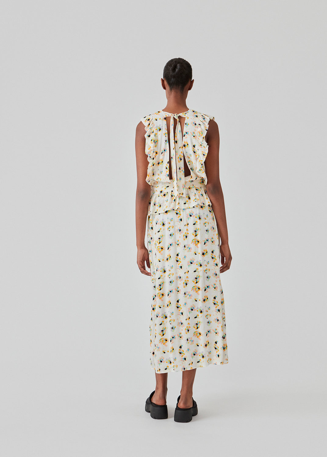 Sleeveless top in the color Flower Sparkles with a high neck and a slight peplum effect. DafneMD print top has ruffled details over the shoulders and at the bottom, along with an opening in the back with ties. The model is 177 cm and wears a size S/36.