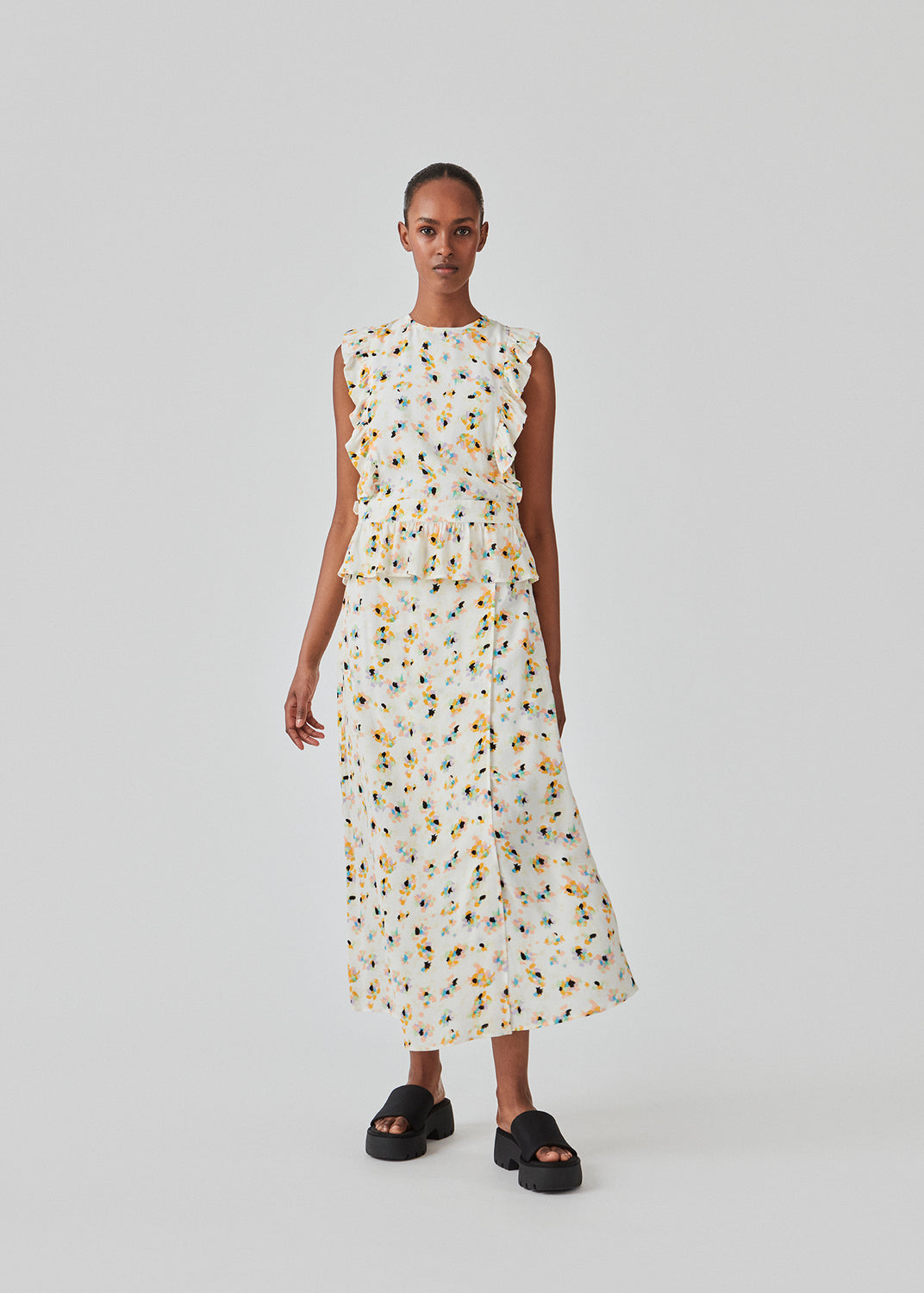 Sleeveless top in the color Flower Sparkles with a high neck and a slight peplum effect. DafneMD print top has ruffled details over the shoulders and at the bottom, along with an opening in the back with ties. The model is 177 cm and wears a size S/36.