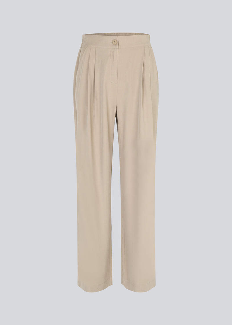 FanyaMD pants in bright beige have a menswear-inspired look with straight, wide legs, a high waist with zip fly and button and elastication in back. Doubble pleat in front and side pockets. The model is 175 cm and wears a size S/36.