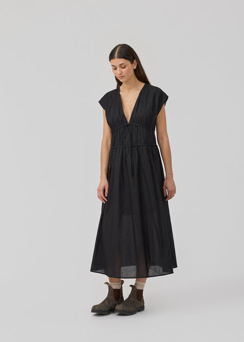 Sleeveless black maxi dress with a loose fit. IndiaMD dress has a deep v-neckline and a tie band at the waist and under the chest. The model is 177 cm and wears a size S/36.