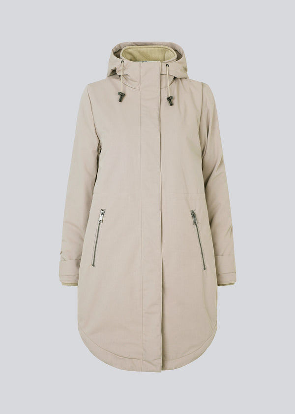 A warm water-repellent winter coat in beige with a high collar and hood. Keller coat has a hidden button and zipper closure at the front, along with zipped pockets. The coat has a relaxed silhouette with an adjustable elastic at the waist. The filling is a sustainable polyester padding with an extra high insulation ability.