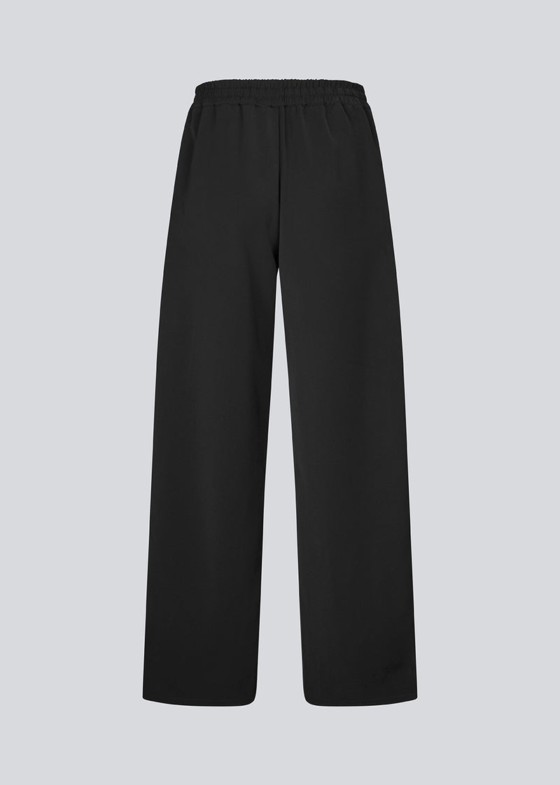 Loose black trousers with wide legs and a relaxed fit. Featuring an elastic waistband with drawstring for a comfortable, adjustable waist. White side stripes add a sporty and modern touch to the otherwise classic design. Perfect for casual everyday wear or more elevated styling – easy to pair with everything from t-shirts to dressy tops.

