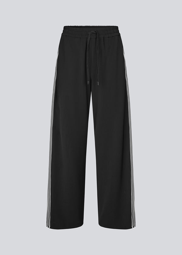 Loose black trousers with wide legs and a relaxed fit. Featuring an elastic waistband with drawstring for a comfortable, adjustable waist. White side stripes add a sporty and modern touch to the otherwise classic design. Perfect for casual everyday wear or more elevated styling – easy to pair with everything from t-shirts to dressy tops.
