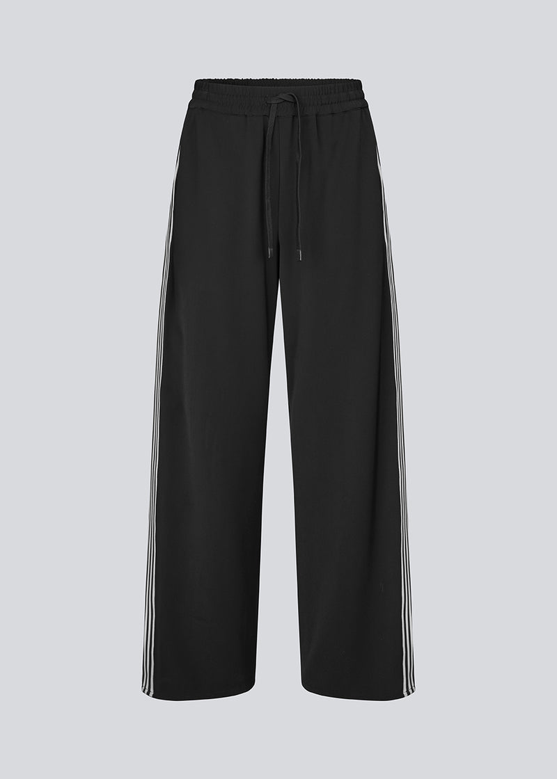 Loose black trousers with wide legs and a relaxed fit. Featuring an elastic waistband with drawstring for a comfortable, adjustable waist. White side stripes add a sporty and modern touch to the otherwise classic design. Perfect for casual everyday wear or more elevated styling – easy to pair with everything from t-shirts to dressy tops.
