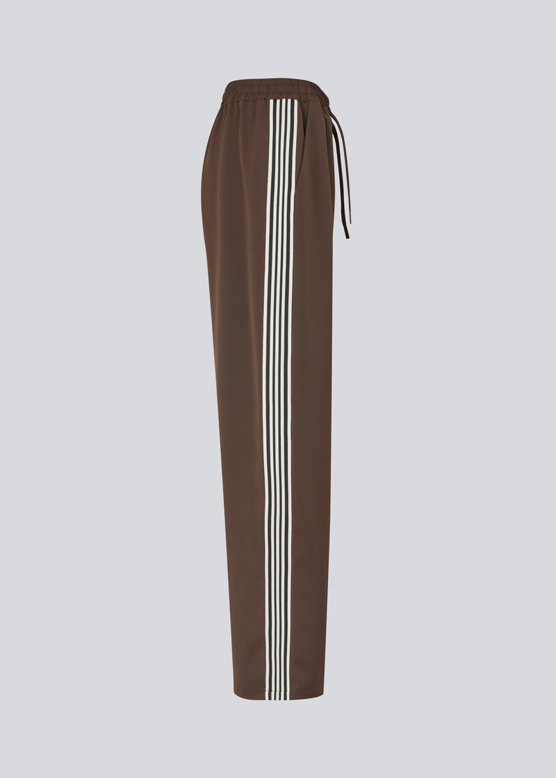 Loose brown trousers with wide legs and a relaxed fit. Featuring an elastic waistband with drawstring for a comfortable, adjustable waist. White side stripes add a sporty and modern touch to the otherwise classic design. Perfect for casual everyday wear or more elevated styling – easy to pair with everything from t-shirts to dressy tops.