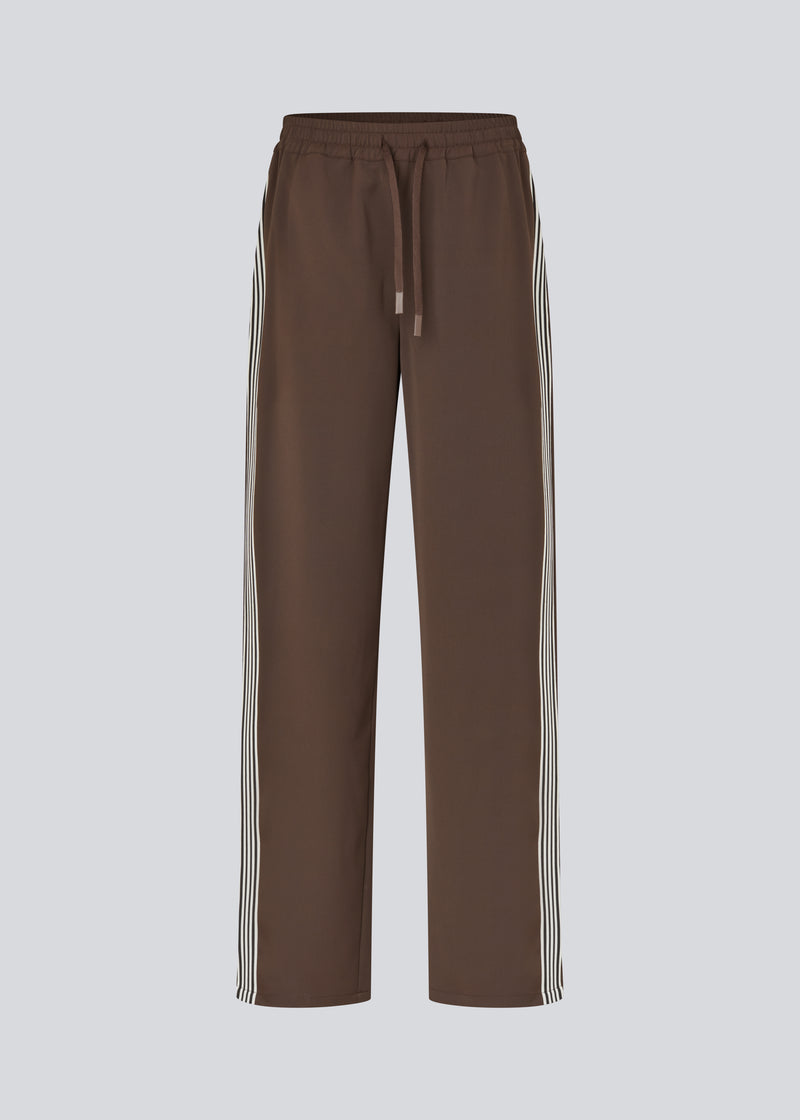 Loose brown trousers with wide legs and a relaxed fit. Featuring an elastic waistband with drawstring for a comfortable, adjustable waist. White side stripes add a sporty and modern touch to the otherwise classic design. Perfect for casual everyday wear or more elevated styling – easy to pair with everything from t-shirts to dressy tops.