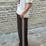Loose brown trousers with wide legs and a relaxed fit. Featuring an elastic waistband with drawstring for a comfortable, adjustable waist. White side stripes add a sporty and modern touch to the otherwise classic design. Perfect for casual everyday wear or more elevated styling – easy to pair with everything from t-shirts to dressy tops.