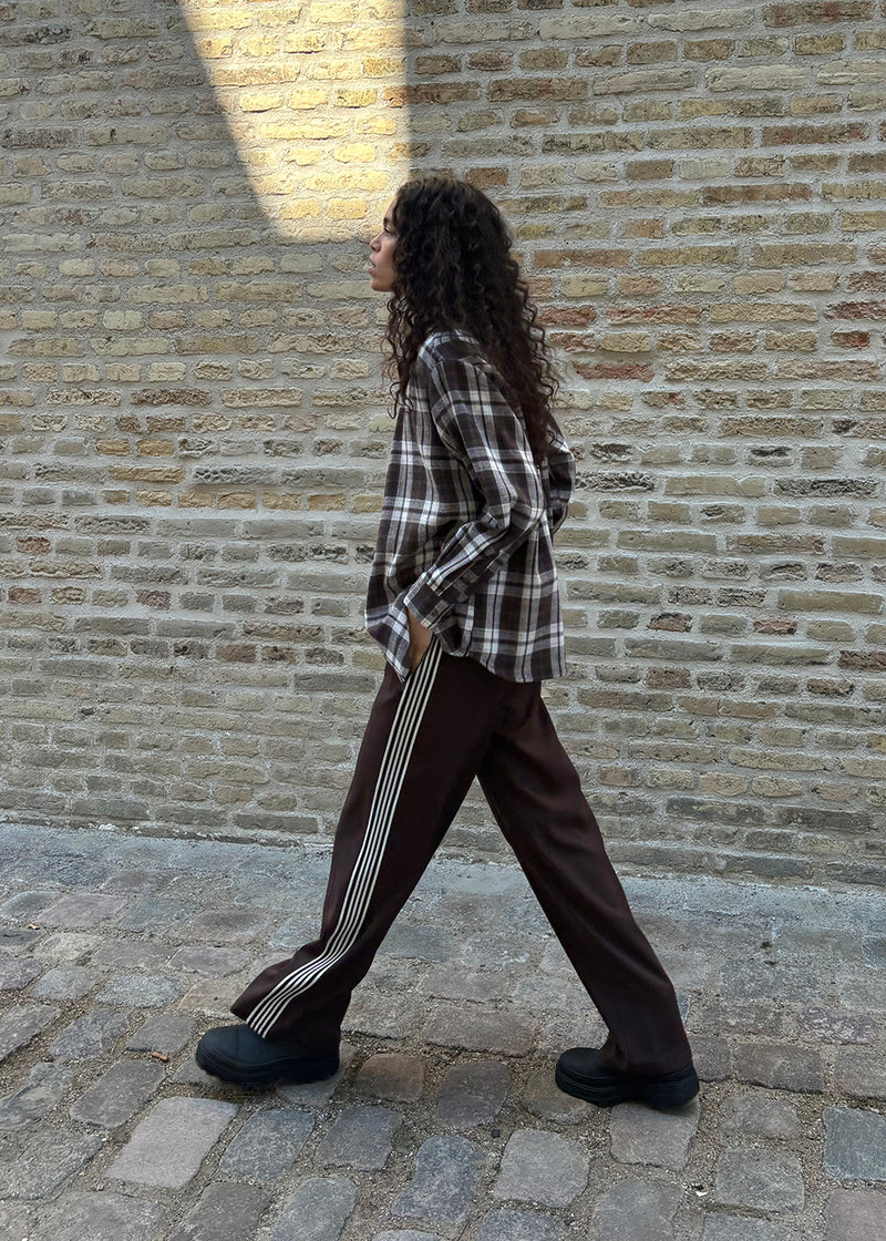 Loose brown trousers with wide legs and a relaxed fit. Featuring an elastic waistband with drawstring for a comfortable, adjustable waist. White side stripes add a sporty and modern touch to the otherwise classic design. Perfect for casual everyday wear or more elevated styling – easy to pair with everything from t-shirts to dressy tops.