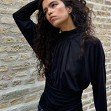 Elegant black blouse featuring a high neckline and long sleeves. Designed with delicate gathered details at the bust and waist to create a flattering silhouette. A subtle button closure at the back of the neck adds a refined and feminine touch. The stretchy fabric ensures a comfortable fit, while the draped design offers timeless elegance. Perfect for both everyday wear and special occasions.
