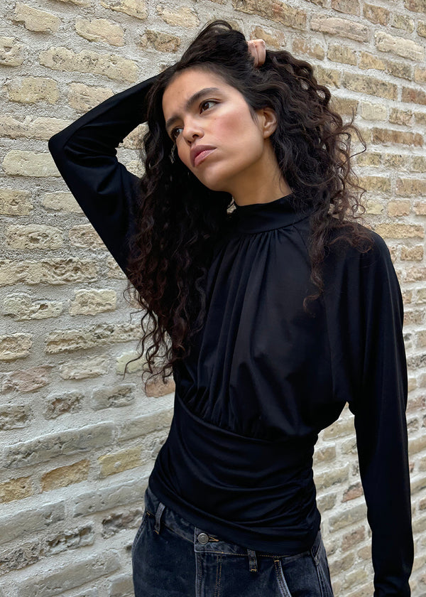 Elegant black blouse featuring a high neckline and long sleeves. Designed with delicate gathered details at the bust and waist to create a flattering silhouette. A subtle button closure at the back of the neck adds a refined and feminine touch. The stretchy fabric ensures a comfortable fit, while the draped design offers timeless elegance. Perfect for both everyday wear and special occasions.
