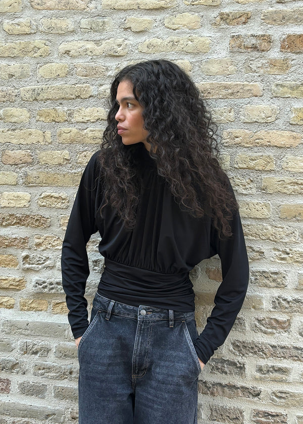 Elegant black blouse featuring a high neckline and long sleeves. Designed with delicate gathered details at the bust and waist to create a flattering silhouette. A subtle button closure at the back of the neck adds a refined and feminine touch. The stretchy fabric ensures a comfortable fit, while the draped design offers timeless elegance. Perfect for both everyday wear and special occasions.