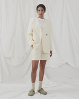 DimeMD blazer is designed in a textured cotton blend with lining. The blazer is collarless but has an asymmetrical wrap detail with a single visible button. The model is 177 cm and wears a size S/36.  Style the blazer with matching shorts: DimeMD shorts.