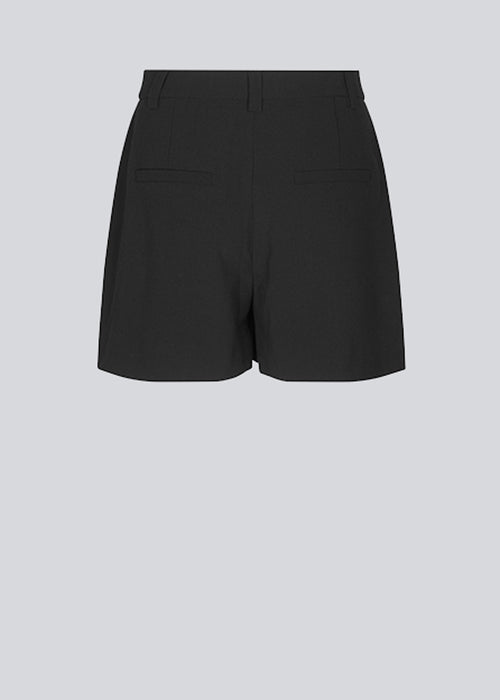 Wide-legged shorts in black with a medium waist. AnkerMD shorts have creases down the front, zip fly and buttons, side pockets, and fake back pockets. The model is 174 cm and wears a size S/36.