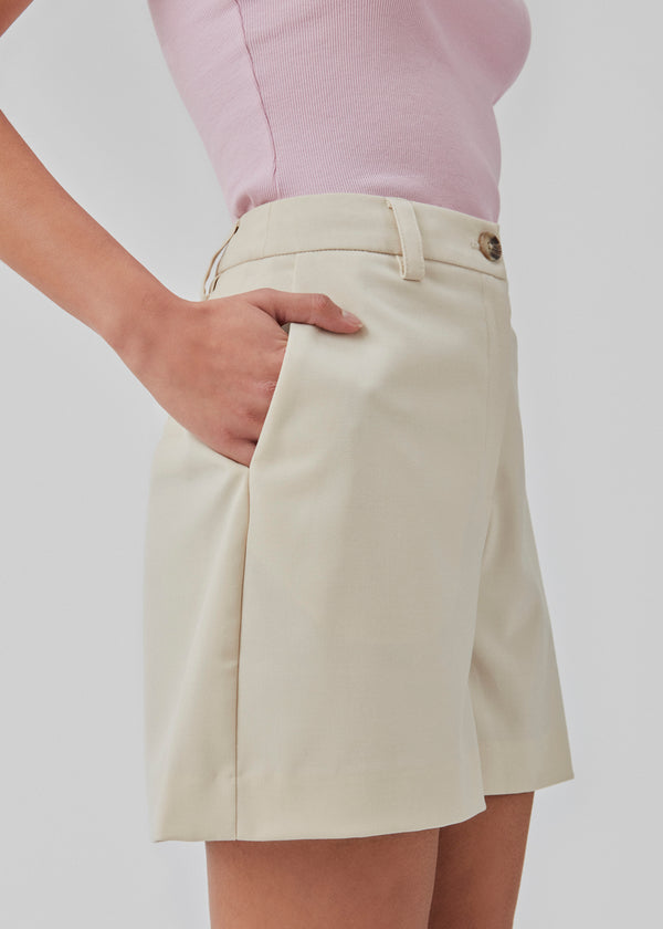 Wide-legged shorts in beige with a medium waist. AnkerMD shorts have creases down the front, zip fly and button, side pockets, and fake back pockets.   The model is 174 cm and wears a size S/36.