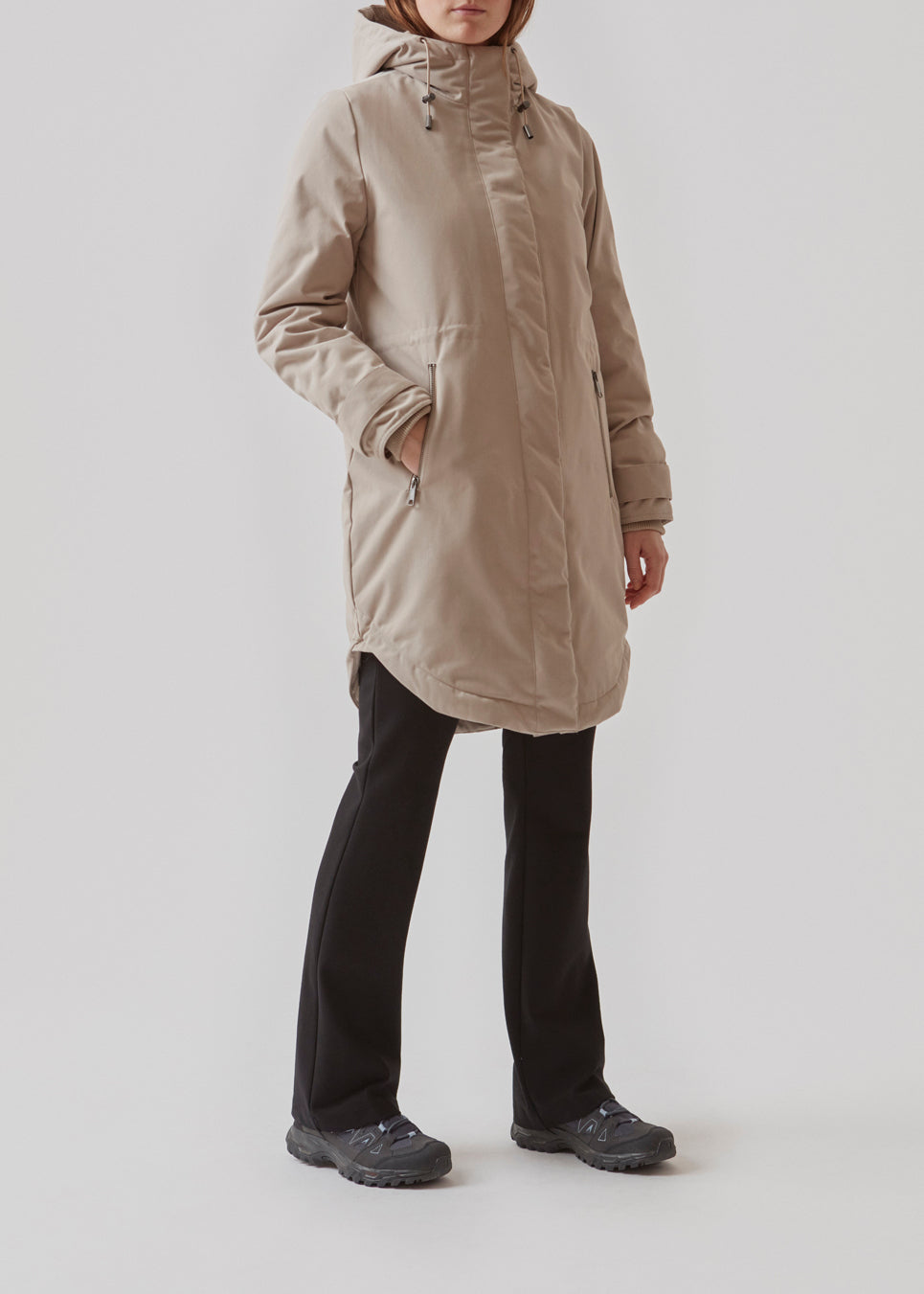 A warm water-repellent winter coat in beige with a high collar and hood. Keller coat has a hidden button and zipper closure at the front, along with zipped pockets. The coat has a relaxed silhouette with an adjustable elastic at the waist. The filling is a sustainable polyester padding with an extra high insulation ability.