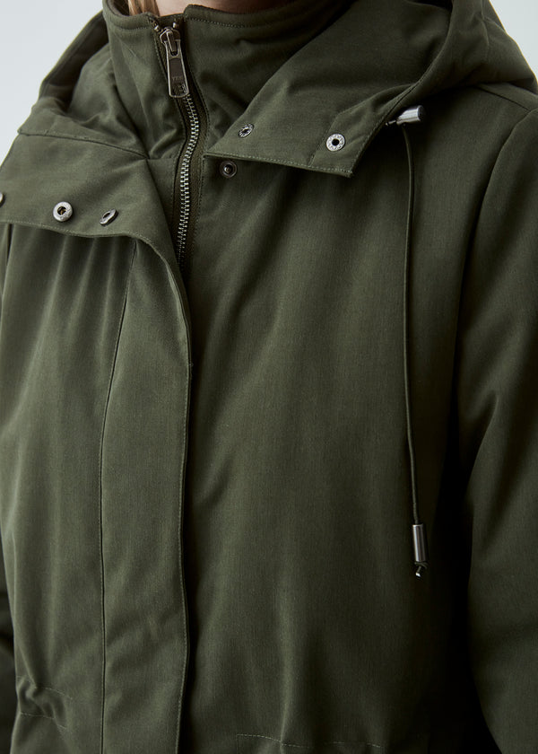 A warm water repellent wintercoat in dark green with a high collar and hood. Keller coat has a hidden button and zipper closure at the front, along with zipped pockets. The coat has a relaxed silhouette with an adjustable elastic at the waist. The filling is a sustainable polyester padding with an extra high insulation ability.