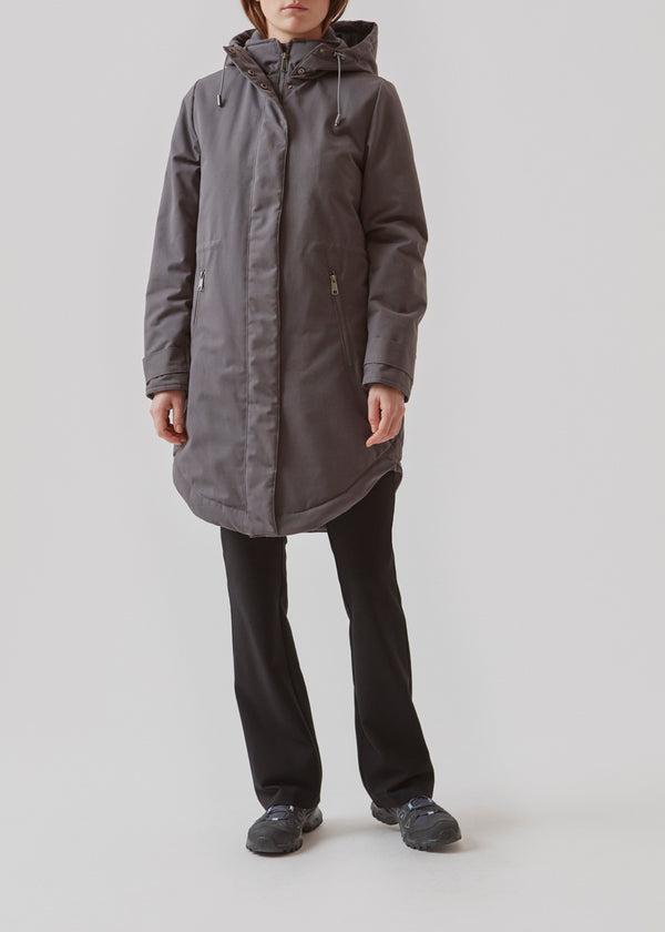 A warm water repellent wintercoat in grey with a high collar and hood. Keller coat has a hidden button and zipper closure at the front, along with zipped pockets. The coat has a relaxed silhuette with an adjustable elastic at the waist. Filling is a sustainable polyester padding with an extra high insulation ability.