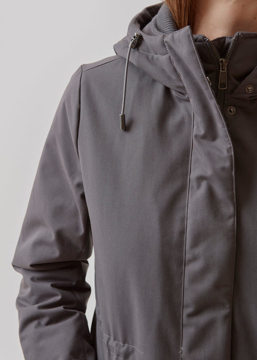 A warm water repellent wintercoat in grey with a high collar and hood. Keller coat has a hidden button and zipper closure at the front, along with zipped pockets. The coat has a relaxed silhuette with an adjustable elastic at the waist. Filling is a sustainable polyester padding with an extra high insulation ability.